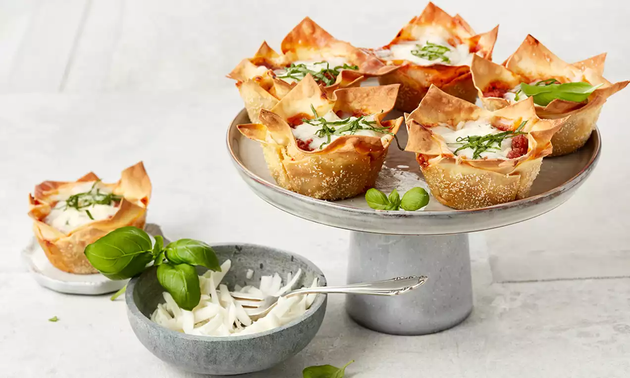 Mini lasagna în foaie aurie de aluat crocant, cu strat de sos roșu, brânză gratinată și frunze proaspete de busuioc pe un suport elegant