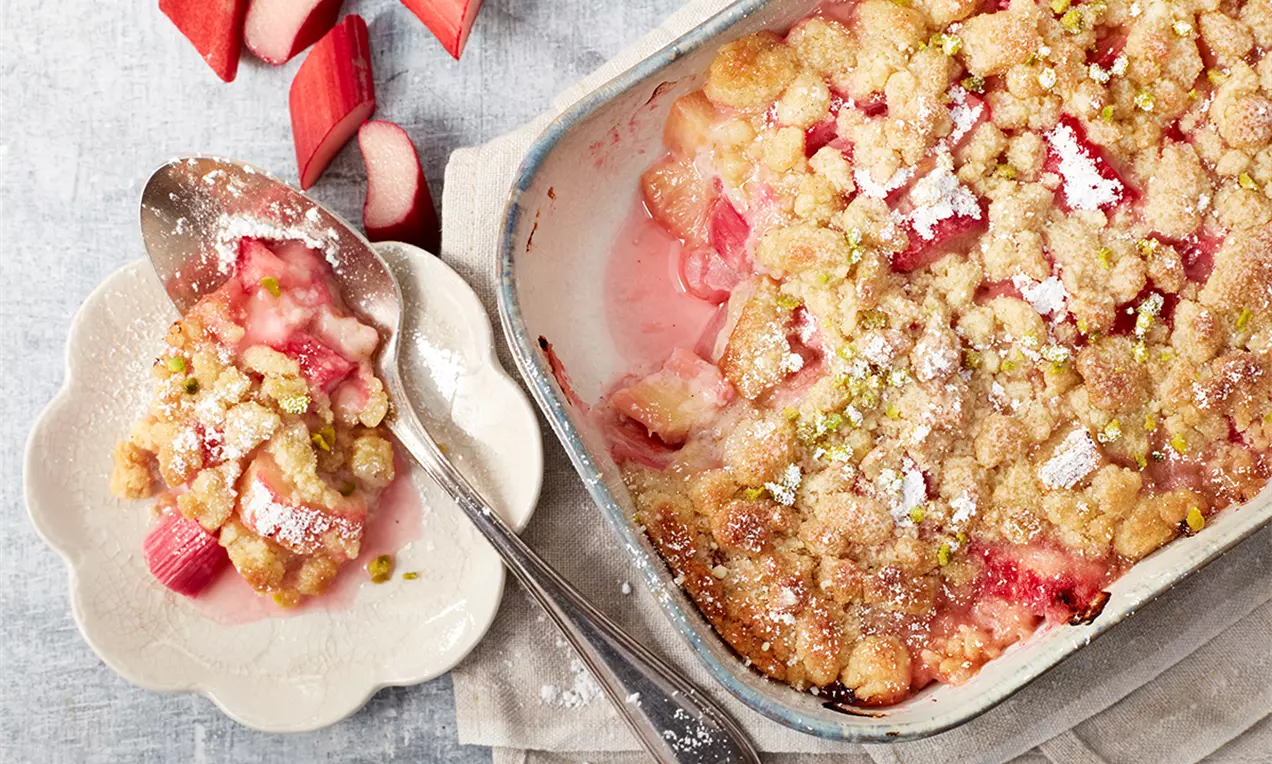 Crumble auriu cu textură crocantă, bucăți roz de rubarbă suculentă, presărat cu fistic și zahăr pudră, servit pe farfurie albă texturată
