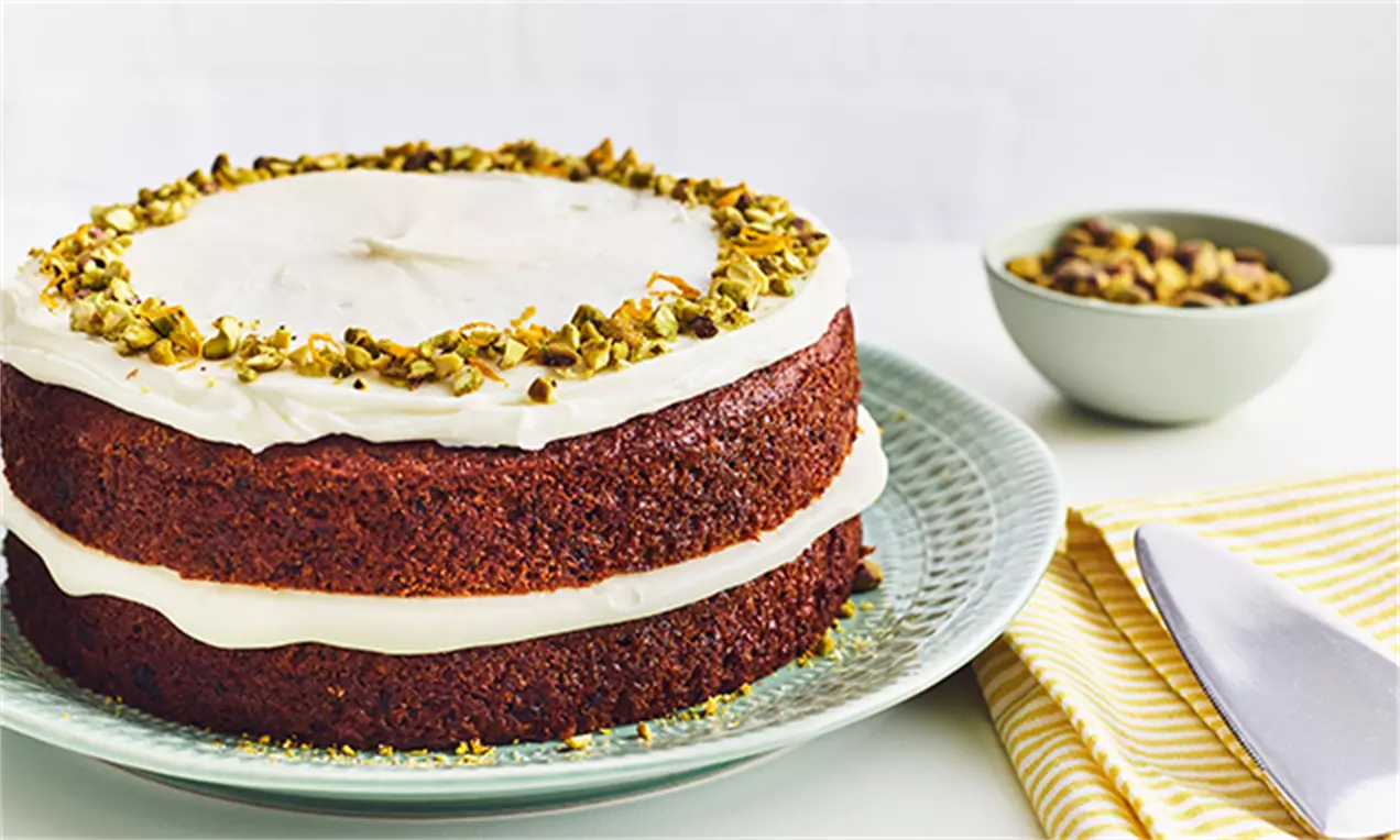 Prăjitură vegană cu morcovi, stratificată cu cremă albă, blat dens și roșiatic, decorată cu migdale pistachio pe o farfurie verde pastel