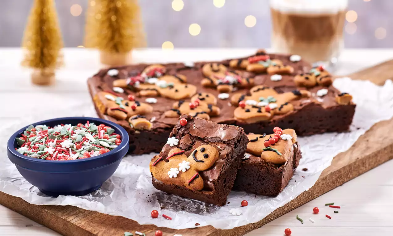 Brownie ciocolatiu dens decorat cu figurine din turtă dulce aurii, fulgi de zahăr albi și ornamente colorate pe hârtie albă creponată