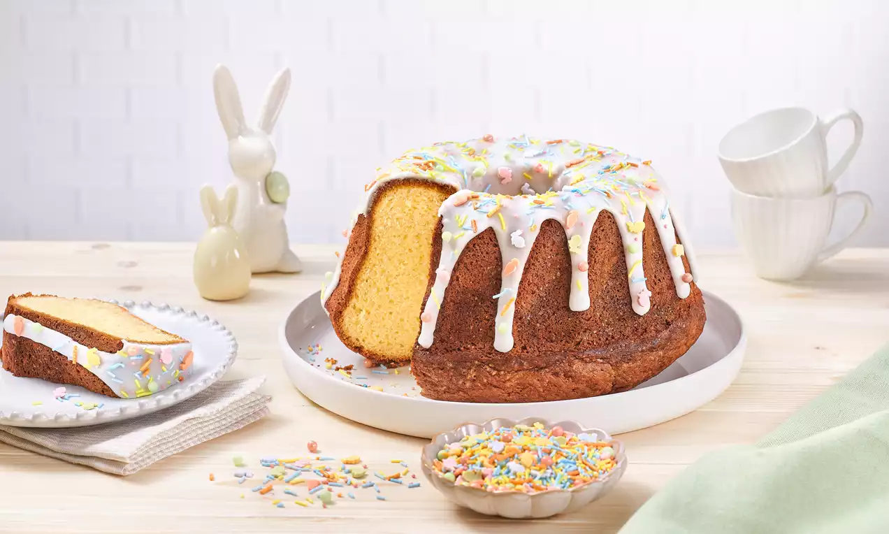 Osterkuchen rotund cu glazură albă lucioasă, decorat cu bombonele colorate pastel pe o farfurie albă lângă un bol și figurine de iepuraș