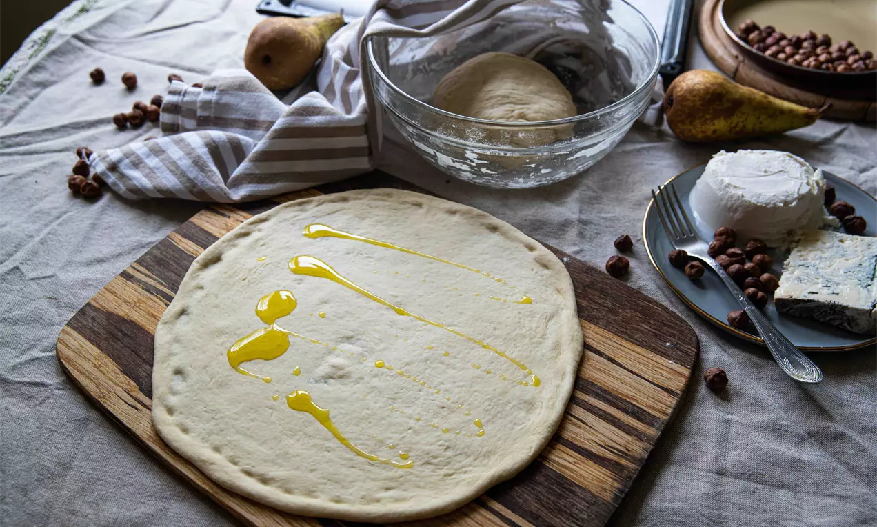 Aluat rotund și neted, deschis la culoare, întins pe un tocător de lemn, stropit cu ulei auriu și înconjurat de ingrediente proaspete