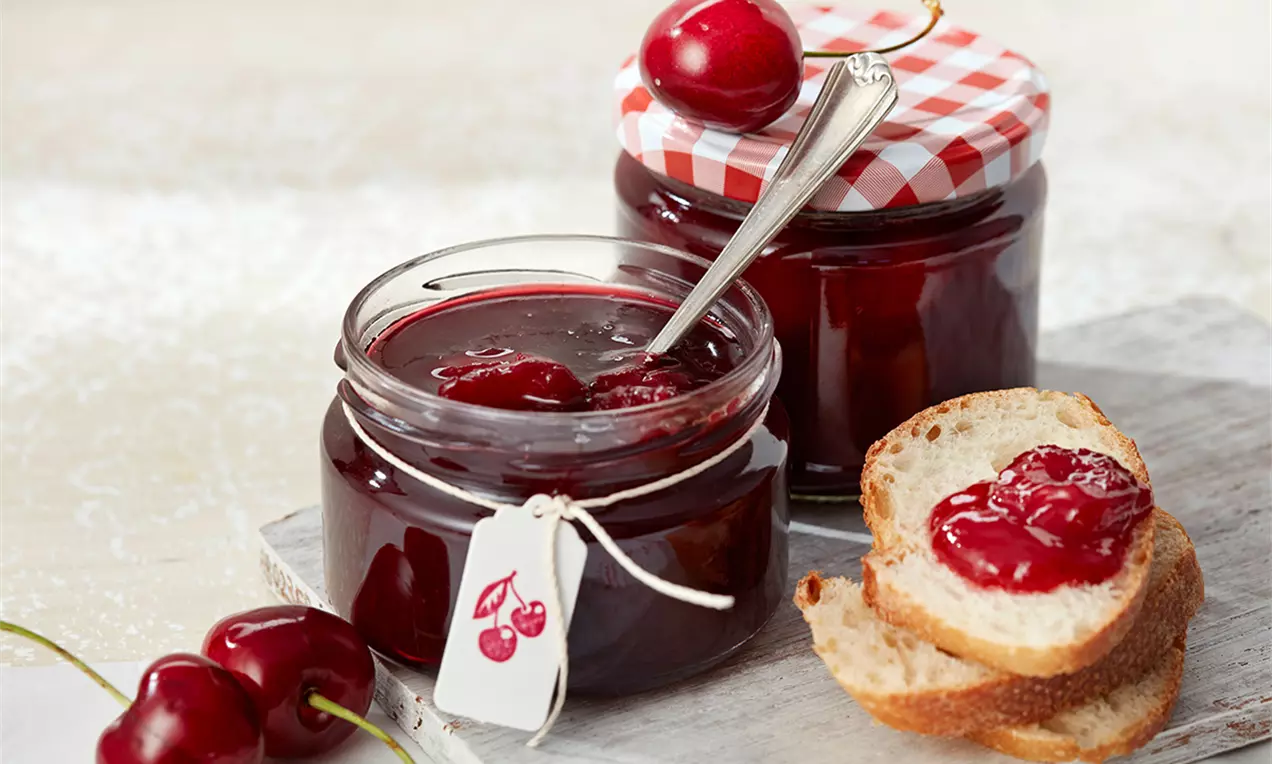 Dulceață roșu-închis de vișine lucioasă în borcane de sticlă, cu capac în carouri și vișine proaspete alături pe o felie de pâine
