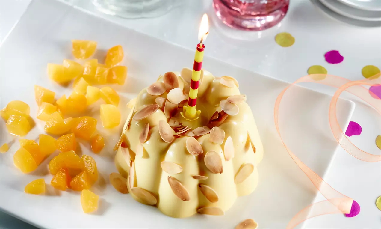 Budincă galben-pal cu textură fină în formă de floare, decorată cu fulgi de migdale și o lumânare, alături de cuburi de mango pe farfurie albă