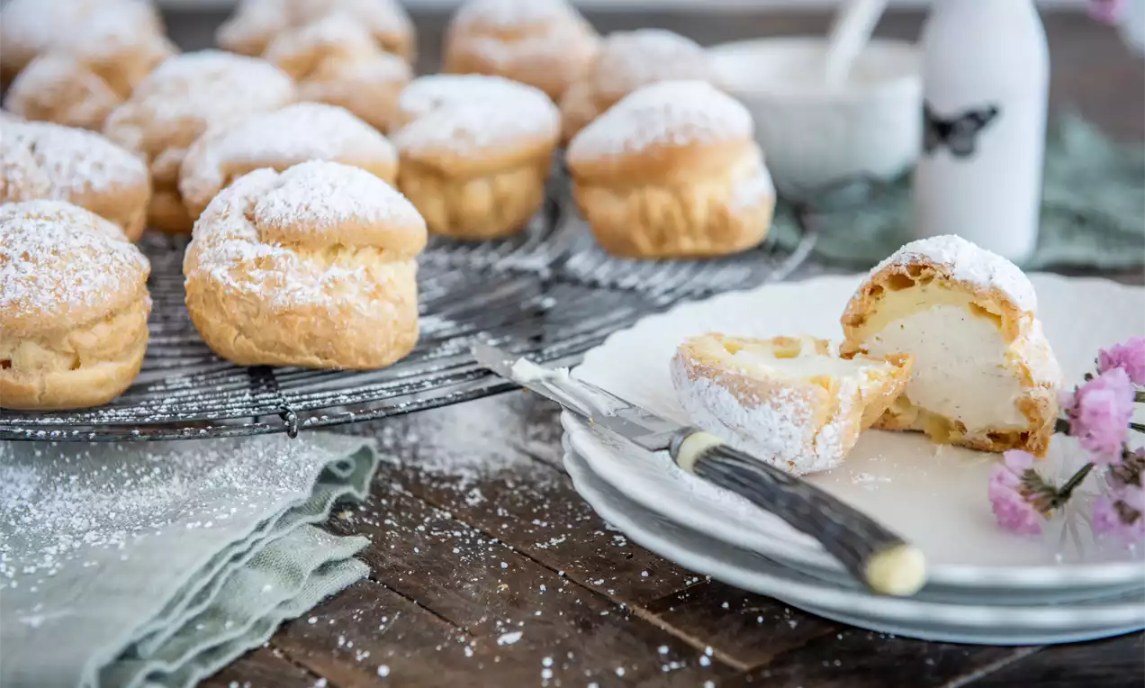 Choux-uri aurii și ușor crocante presărate cu zahăr pudră alb, cu umplutură cremoasă de vanilie, așezate pe un grilaj metalic și o farfurie albă