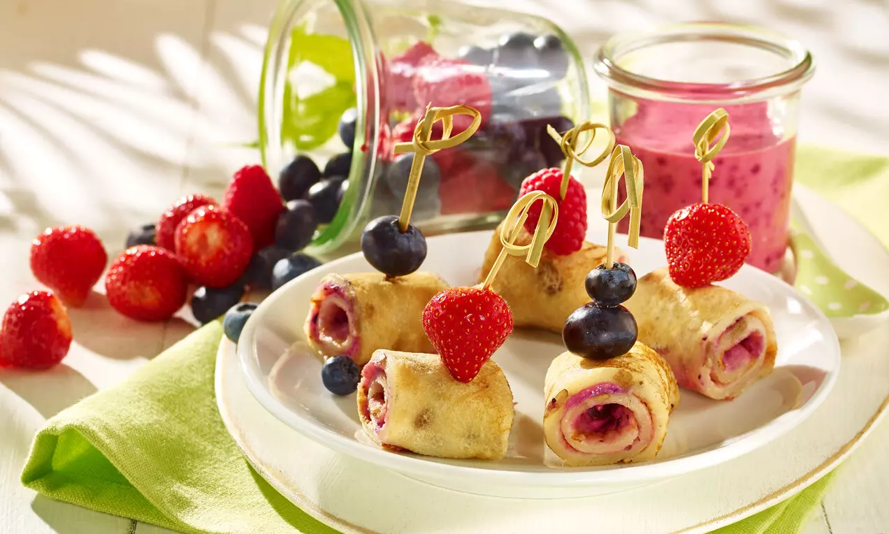 Clătite vegane aurii rulate, umplute cu fructe colorate, prinse cu frigărui și decorate cu căpșuni și afine pe o farfurie albă