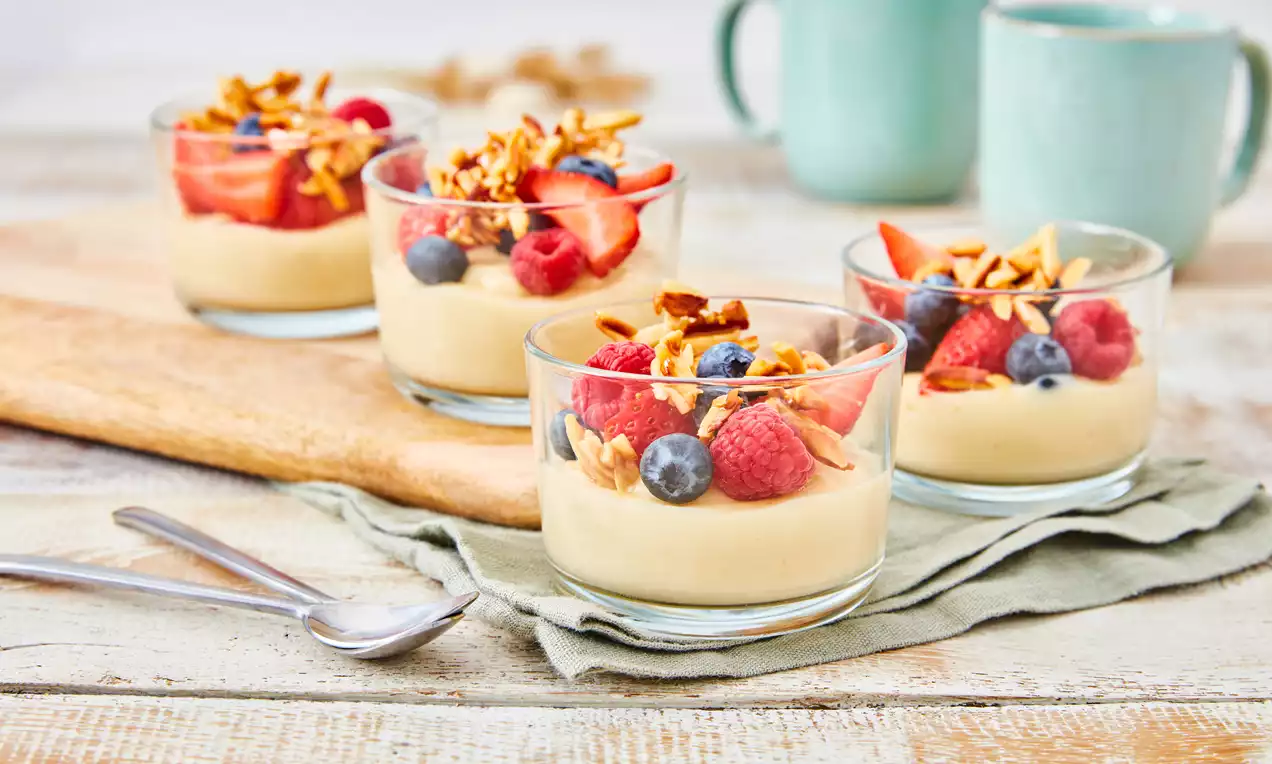Pudding cremos galben pal în pahare rotunde, decorat cu fructe roșii și albastre, granola aurie pe deasupra, așezat pe o lavetă verde pal