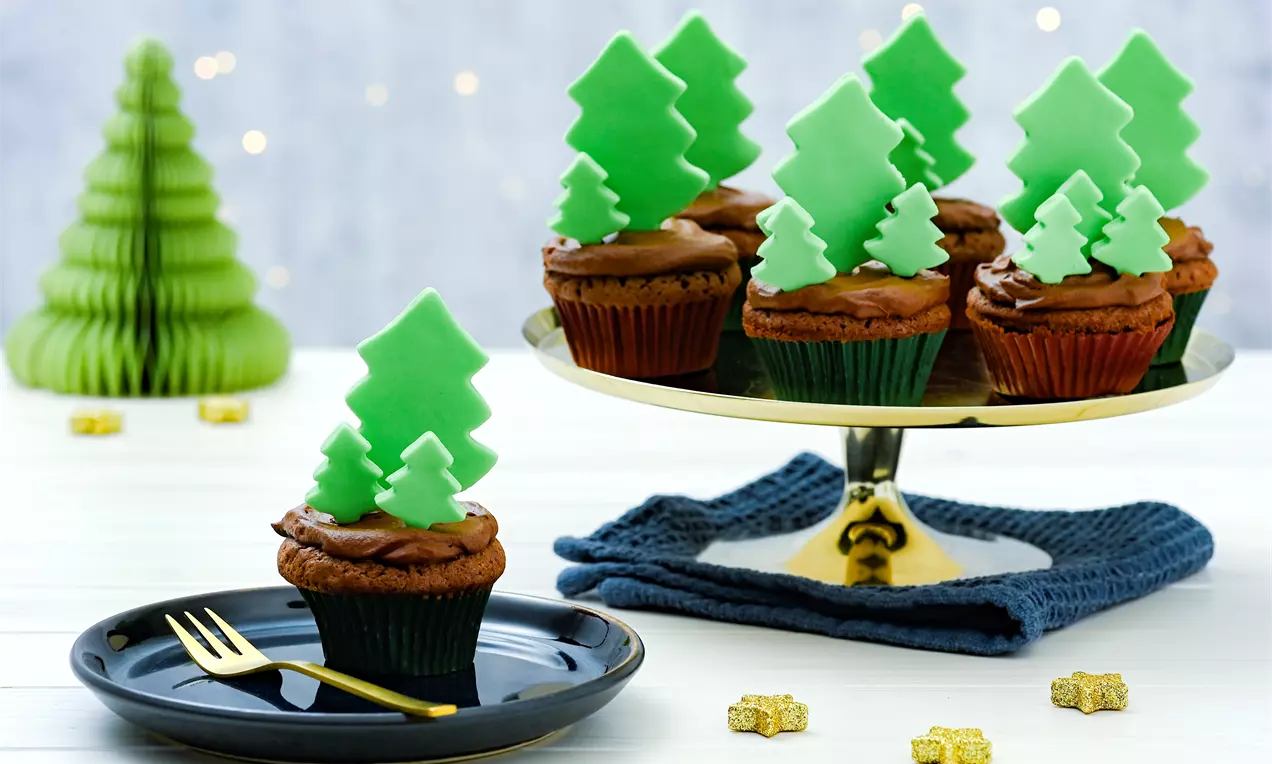 Brioșe pufoase decorate cu cremă de ciocolată și brăduți verzi din fondant, aranjate pe un platou auriu lângă decorațiuni festive