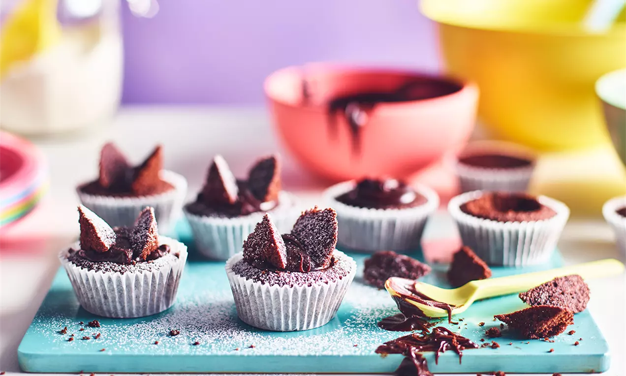 Cupcakes cu blat de ciocolată moale, decorate cu aripi de fluture din bucăți de prăjitură, presărate cu zahăr pudră, pe un fundal pastelat