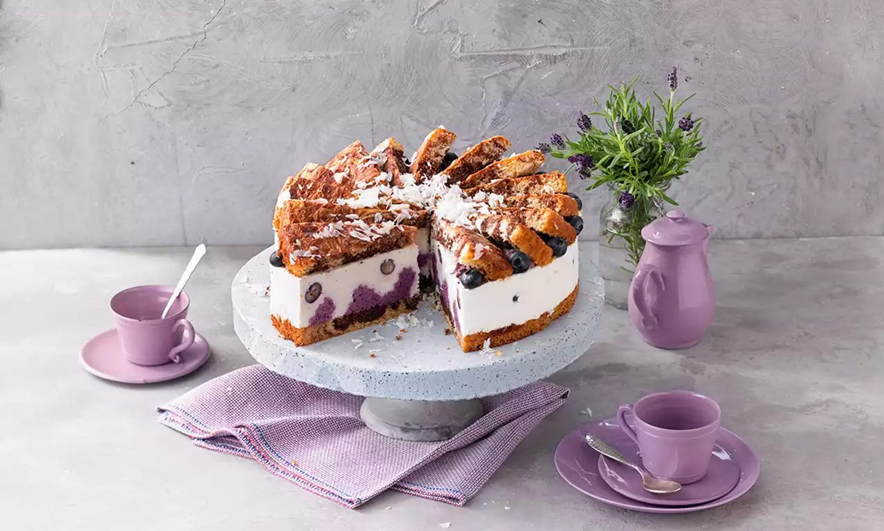 Tort cu mousse de afine pe un platou gri, cu strat alb cremos și afine întregi, acoperit cu blat aurit și fulgi de cocos