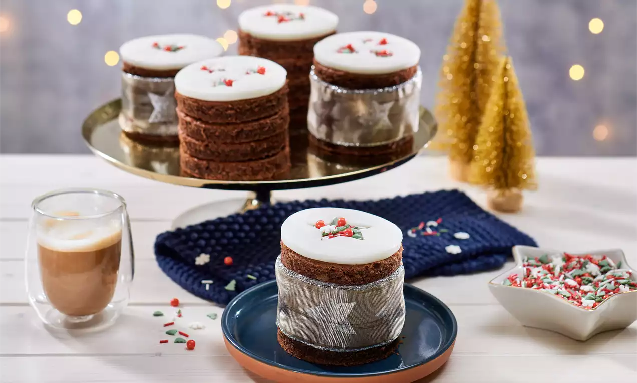 Mini-prajituri rotunde cu blat de cacao maro, glazură albă decorată festiv și panglică argintie, așezate pe platou și farfurii