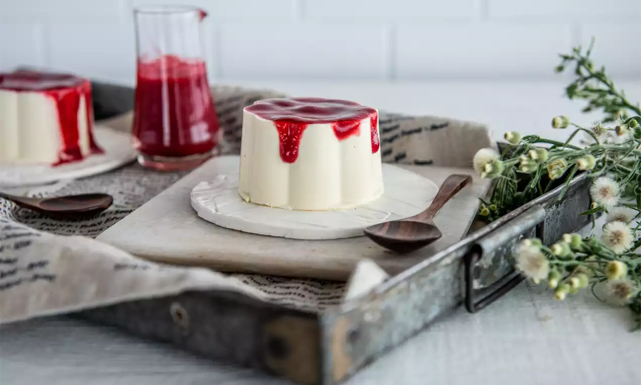 Panna Cotta alb-crem lucioasă, netedă și cilindrică, acoperită cu sos roșu intens, pe o farfurie albă, alături de flori mici și o lingură de lemn
