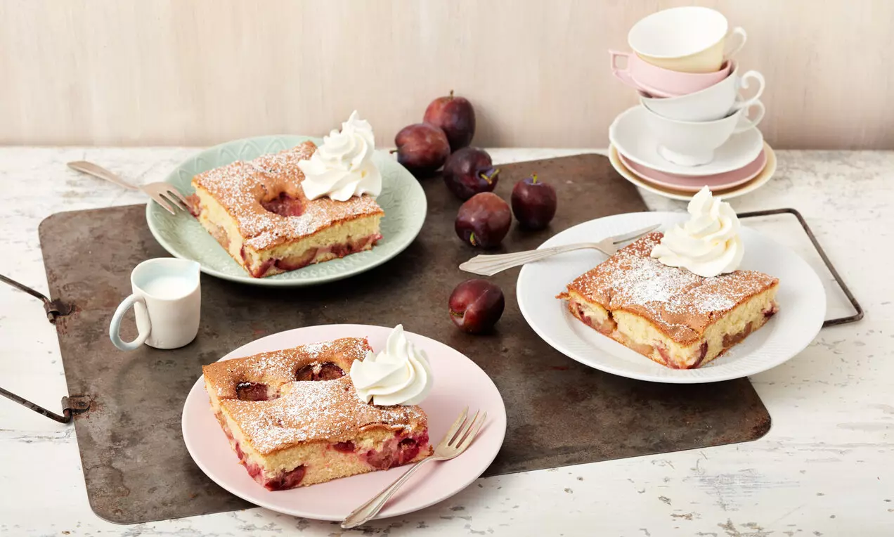 Prăjitură rumenită cu prune roșii coapte, presărată cu zahăr pudră și decorată cu moturi pufoase de frișcă pe farfurii pastelate