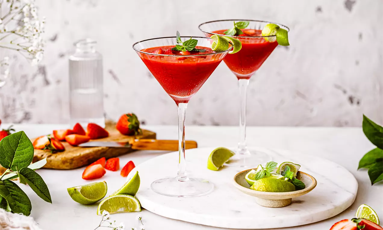 Daiquiri cu căpșuni de un roșu intens servit în cupe de cocktail, decorat cu felii de lime și frunze proaspete de mentă pe un fundal luminos