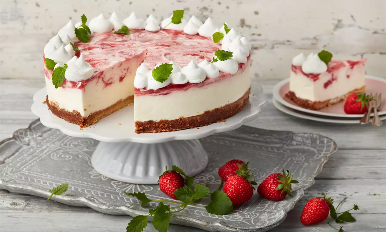 Tort cu strat cremos alb și vârtejuri roz de căpșuni pe blat de biscuiți maro, decorat cu moturi de frișcă și frunze verzi, pe un platou alb