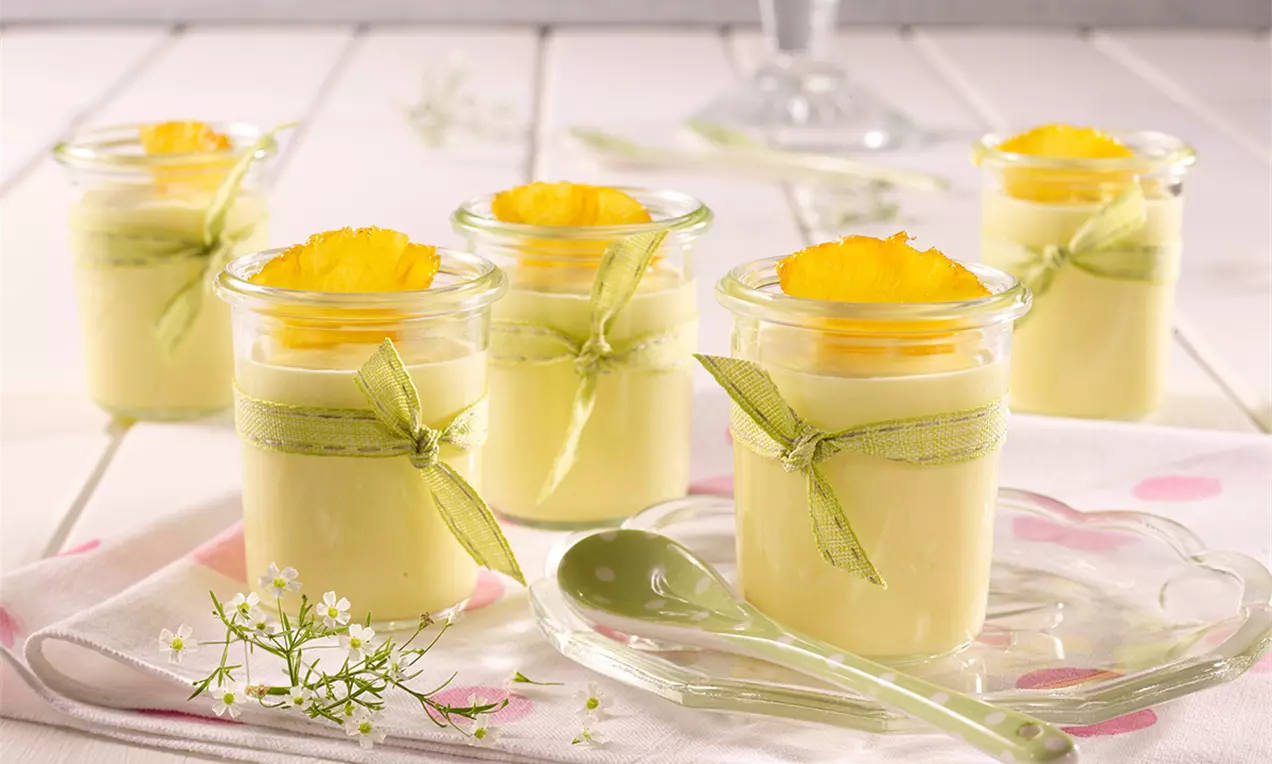 Mousse aerat galben-portocaliu în pahare de sticlă cu panglică verde, decorat cu felii de mango pe o masă albă cu flori delicate