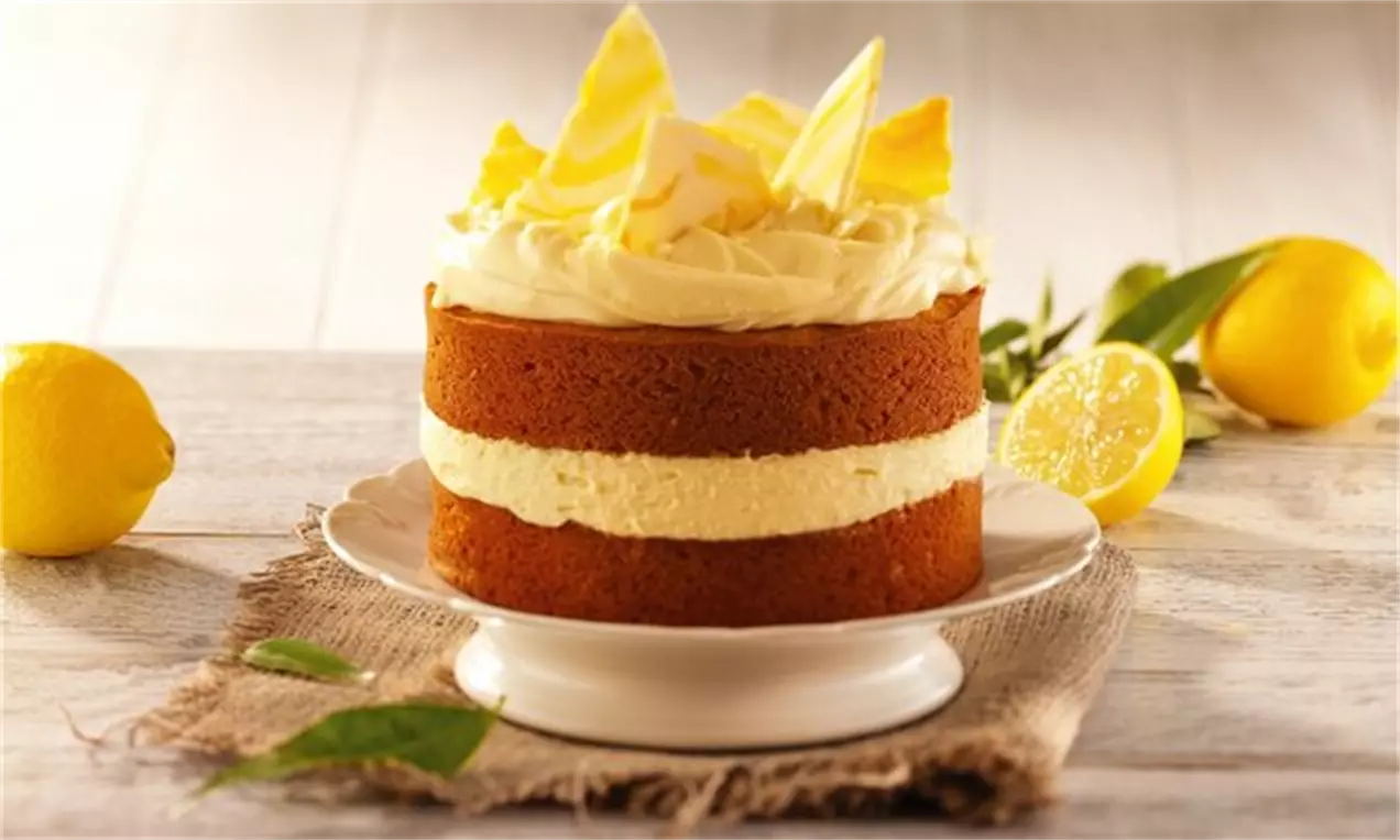 Tort de cheesecake înalt cu straturi aurii de blat pufos, cremă fină de lămâie și ciocolată albă răzuită, pe un platou alb și decorat cu lămâi și frunze