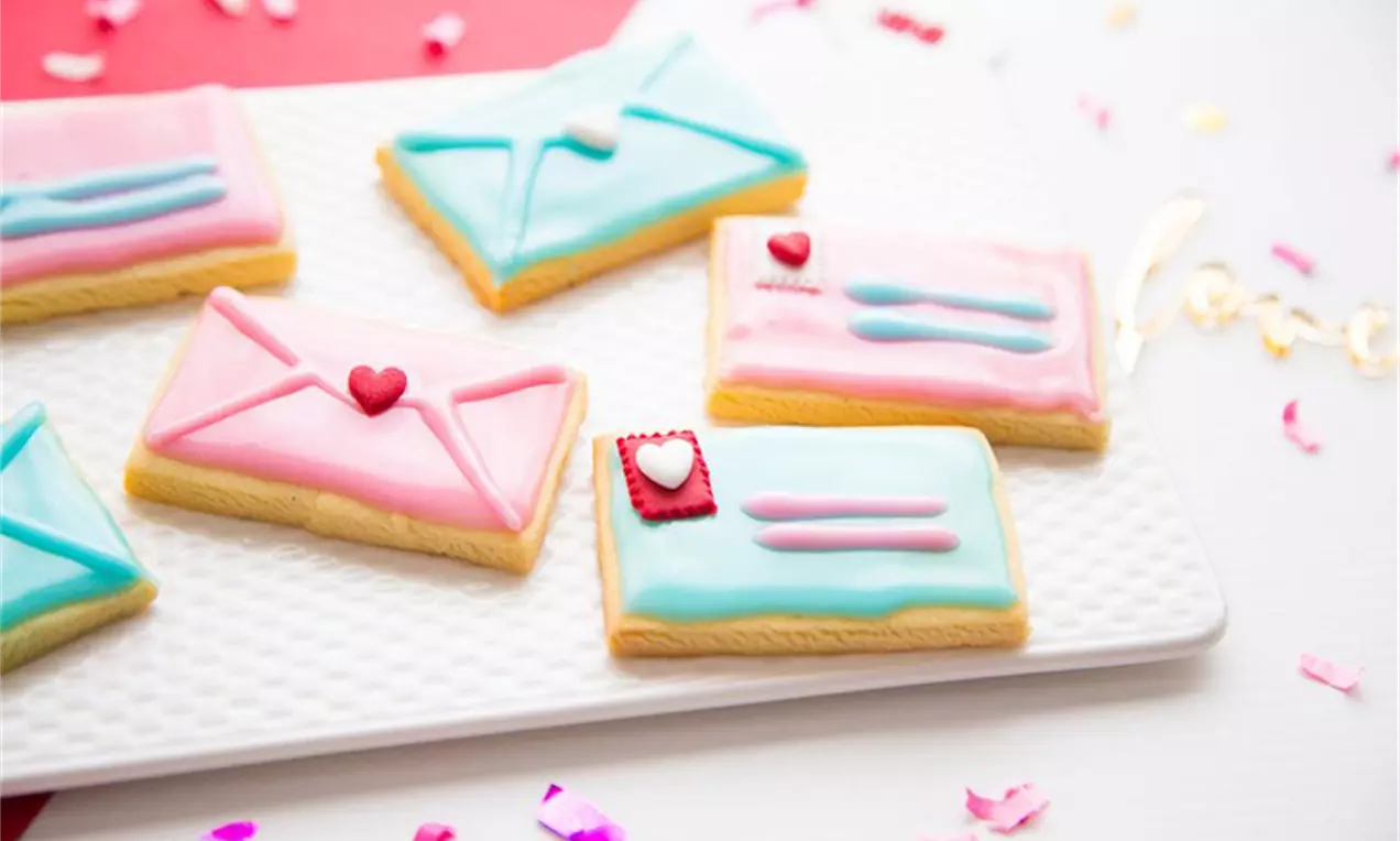 Biscuiți cu unt aurii, decorați cu glazură roz și albastru pastel, în formă de plicuri și scrisori delicate, pe o farfurie albă texturată