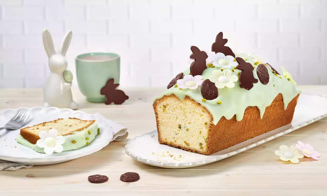 Chec pufos dreptunghiular acoperit cu glazură verde pastel, decorat cu flori din zahăr și iepurași de ciocolată pe un platou alb