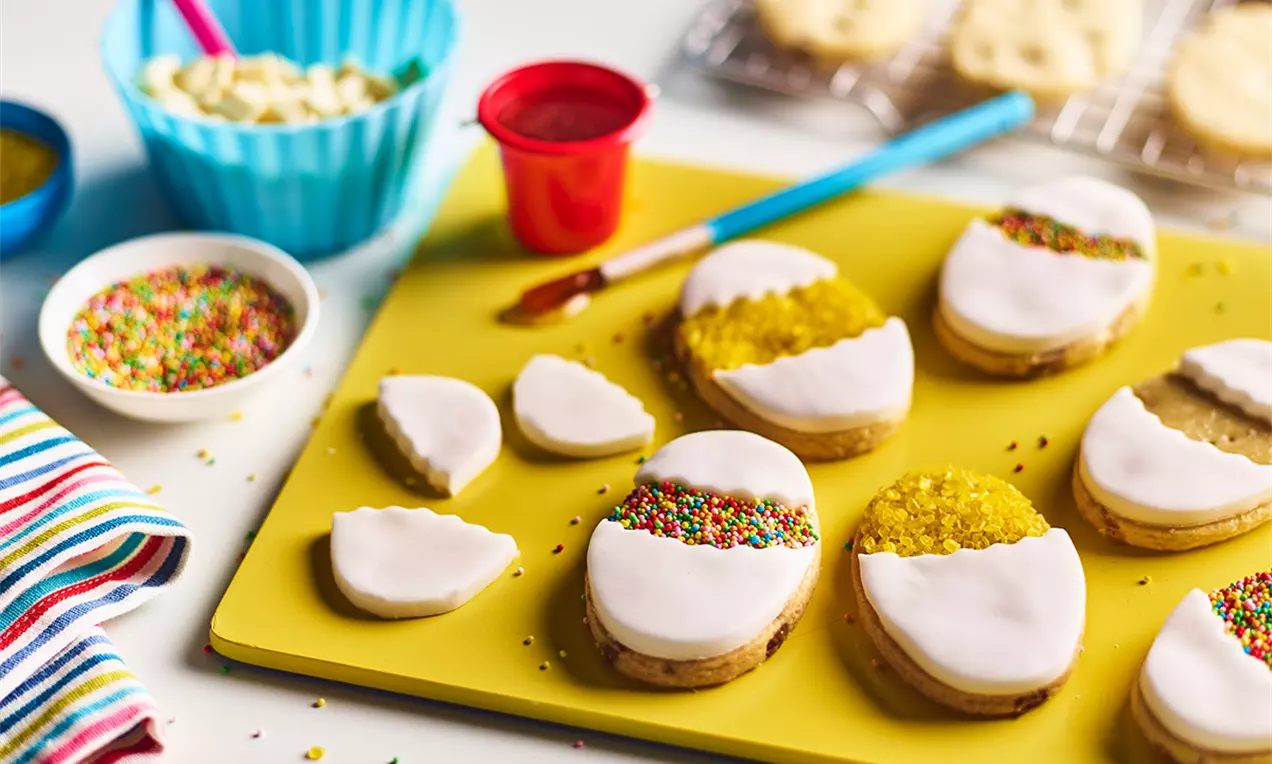 Biscuiți aurii în formă de ouă glazurate cu alb și decor pastelat de zahăr colorat, așezați pe un platou galben viu, înconjurați de garnituri și ustensile pentru ornat