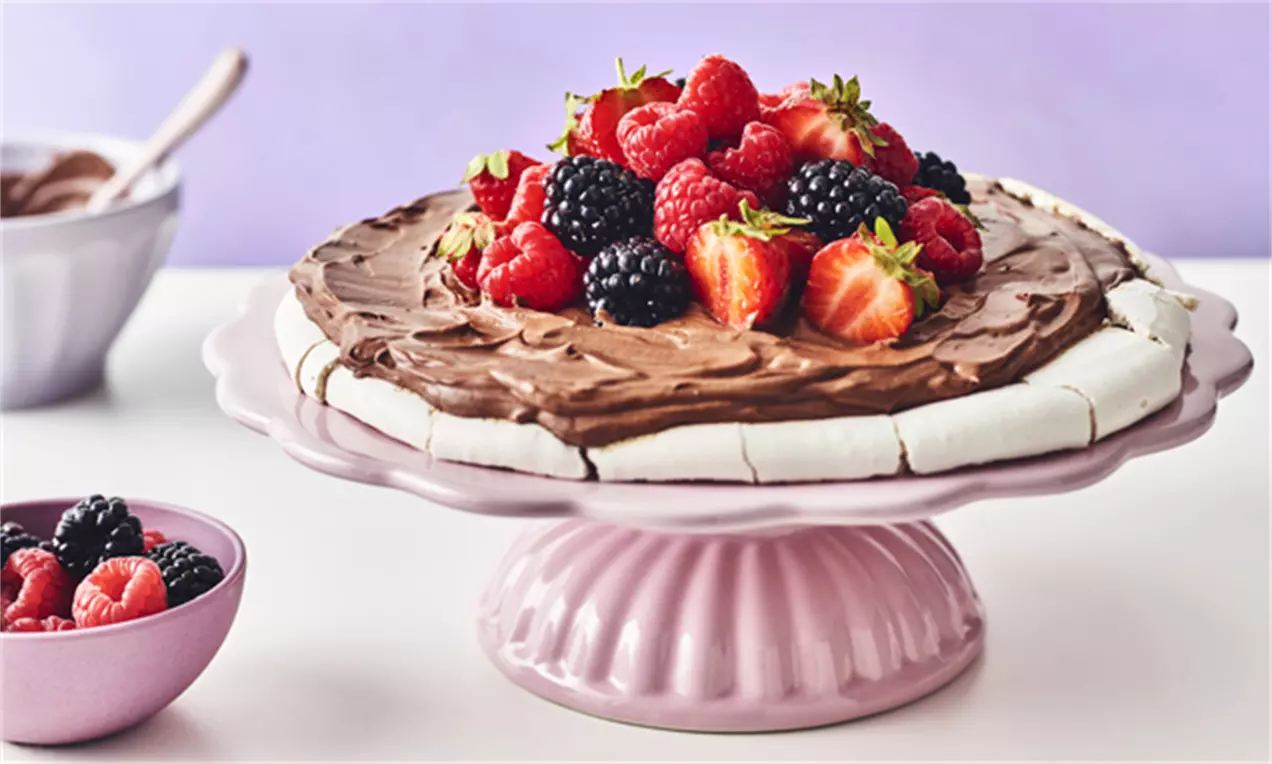 Pavlova vegană albă, aerată, acoperită cu cremă fină de ciocolată și decorată cu fructe roșii și negre proaspete pe un platou roz