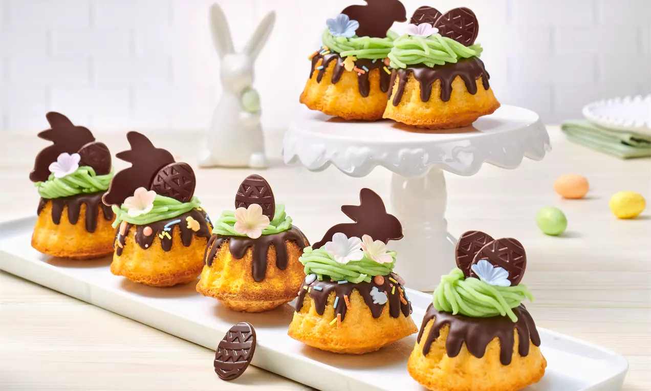 Mini-gugulufuri aurii cu glazură de ciocolată, decor verde de cremă, flori pastelate și figurine din ciocolată așezate pe platouri albe