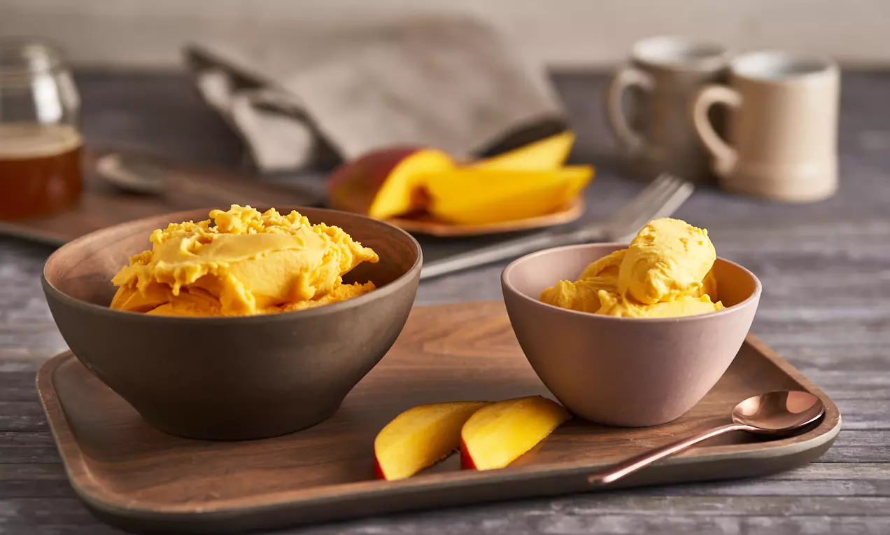 Înghețată vegană de mango galben-intens, cremoasă, servită în boluri mate pe o tavă de lemn, alături de felii proaspete de mango