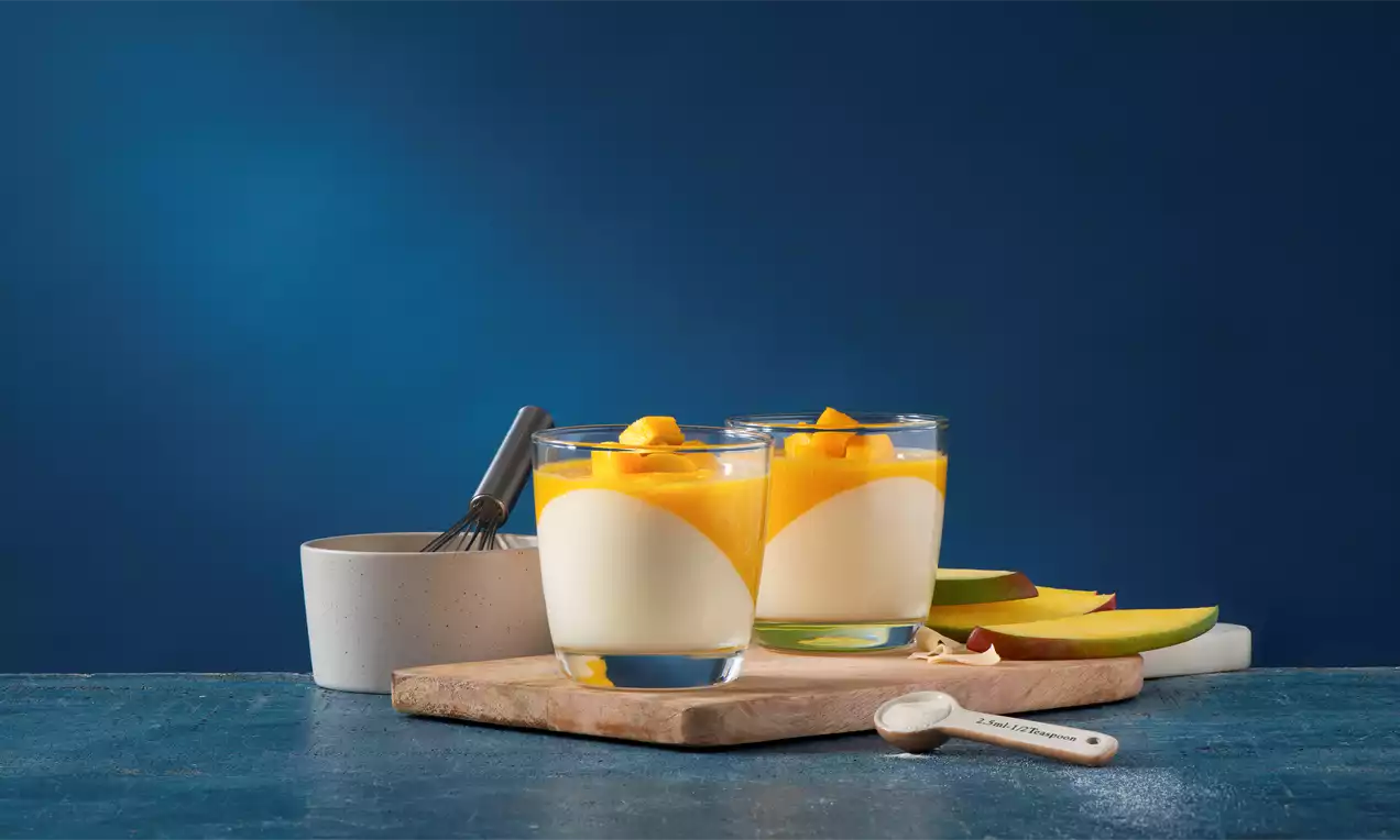 Jeleu alb cu vanilie cremos în pahare transparente, decorat cu mango portocaliu vibrant pe un fundal albastru și blat de lemn