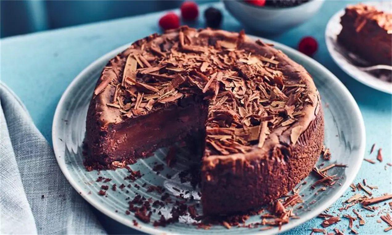 Cheesecake rotund cu suprafață lucioasă de ciocolată, decorat cu fulgi de ciocolată, așezat pe o farfurie albă, cu fructe în fundal
