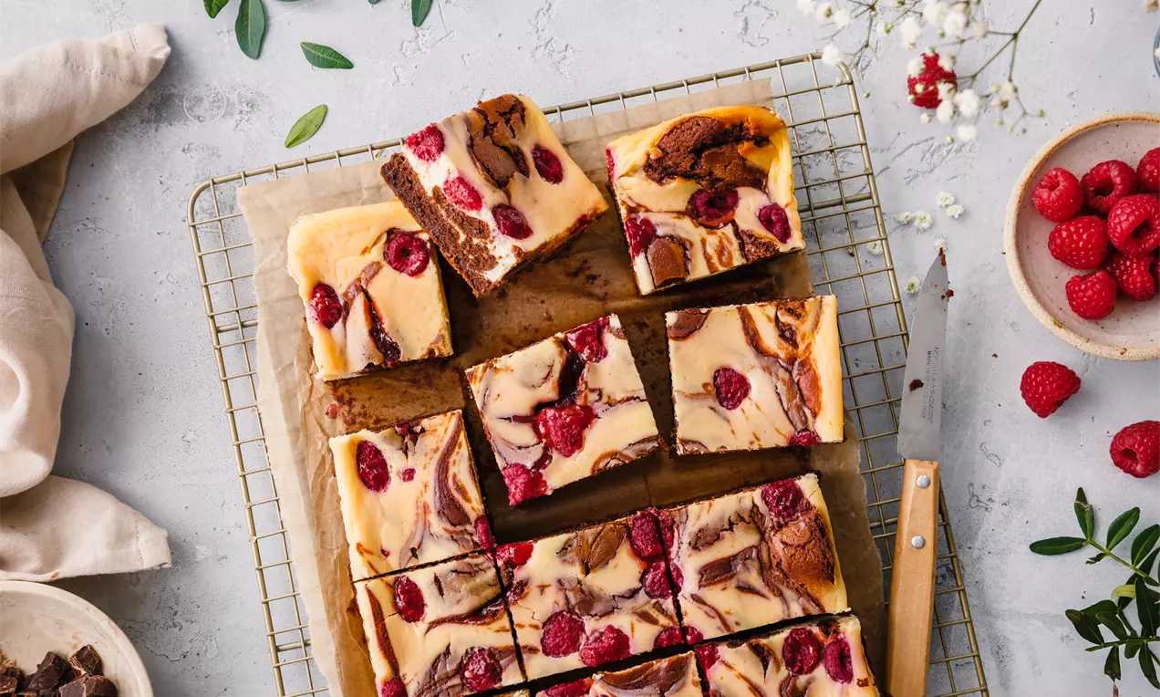 Brownie cu strat cremos de cheesecake vanilat, marmorat cu ciocolată și presărat cu zmeură, tăiat în pătrate și așezat pe un grătar de răcire