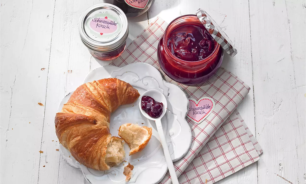 Dulceață roșu-rubiniu cu bucăți lucioase de cireșe într-un borcan de sticlă deschis, lângă un croissant auriu pe o farfurie albă și o linguriță
