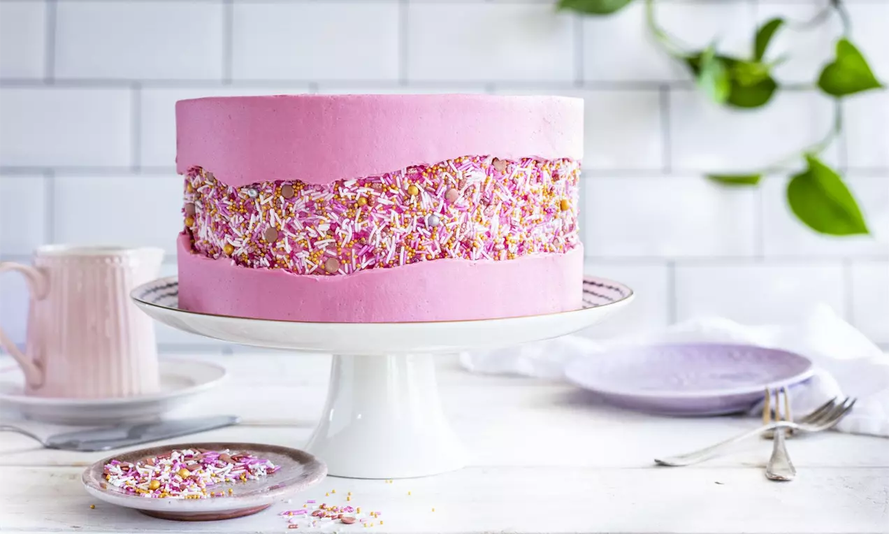 Tort roz decorat cu cremă netedă de unt, ornamente strălucitoare din fulgi colorati în centru, pe un platou alb elegant