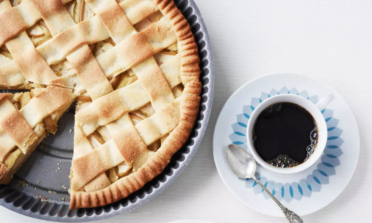 Plăcintă americană aurie cu mere, crusta împletită uniform și crocantă, feliată lângă o ceașcă de cafea neagră pe farfurie decorată