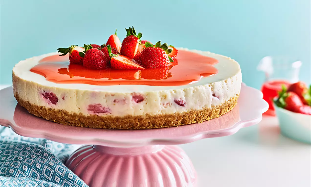 Cheesecake cremos alb cu fragmente de căpșuni și blat auriu, acoperit cu un strat subțire de jeleu roșu și căpșuni proaspete în vârf