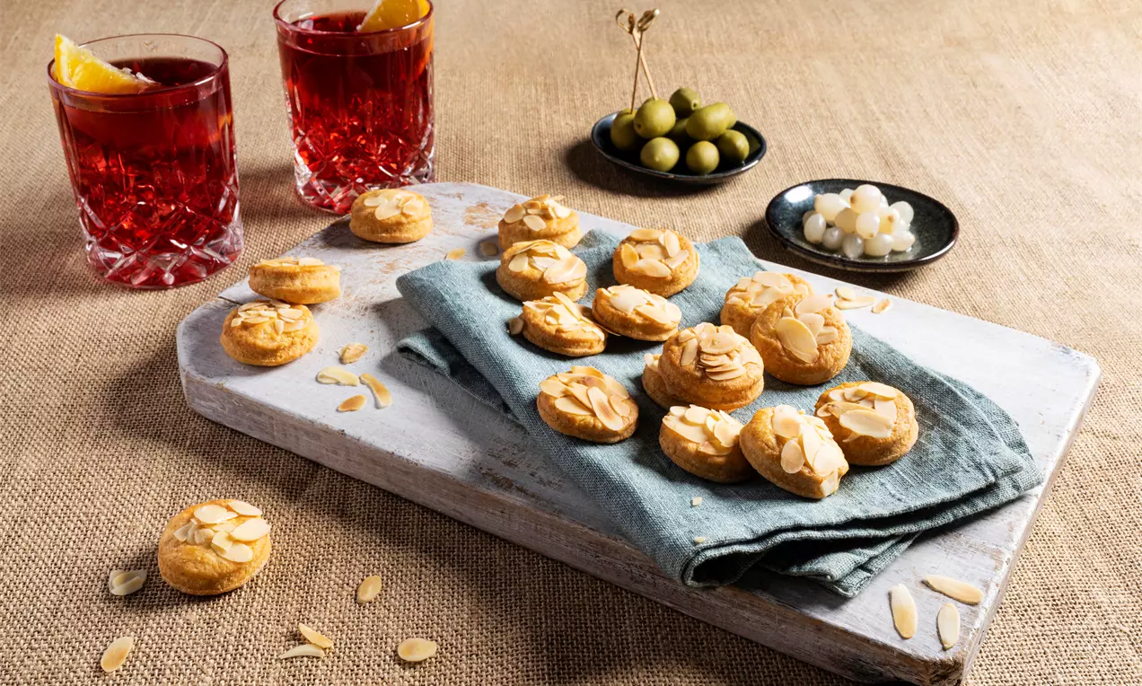 Biscuiți aurii, rotunzi și fragili, presărați cu fulgi de migdale, așezați pe un șervet albastru pe o tăblie albă, însoțiți de două pahare cu băutură roșie, măsline și ceapă murată.