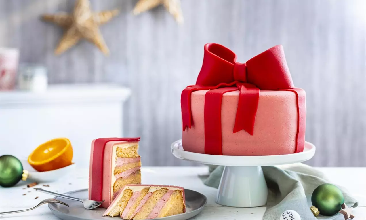 Tort decorat cu fondant roz și panglică roșie de cadou, pe platou alb, alături de felii ce dezvăluie straturi de cremă roz și blat auriu