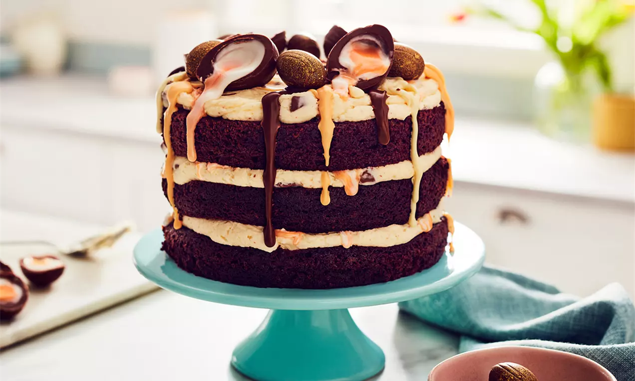 Tort în straturi de blat de cacao intens, cu cremă galben-pastel de ouă și decor din ouă de ciocolată și firicele glazurate pe un platou turcoaz