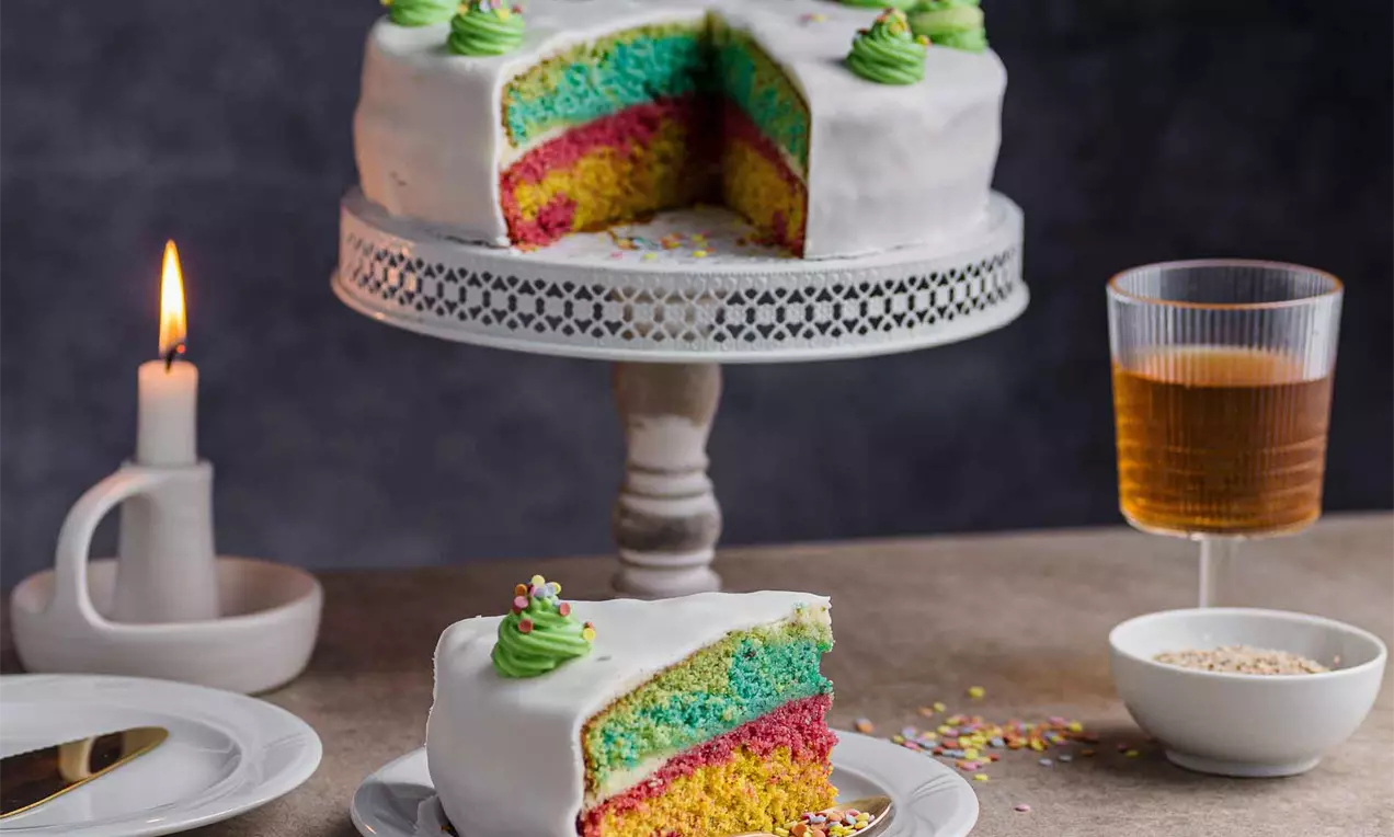 Tort cu straturi viu colorate în nuanțe de galben, roz și albastru, acoperit cu glazură albă și moturi de cremă verde, servit pe suport și farfurie albă.