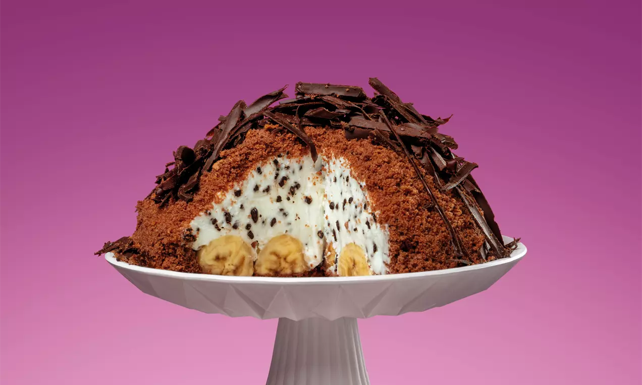 Desert în formă de cupolă acoperit cu firimituri de blat de ciocolată, umplutură albă cremoasă cu bucăți de banană și decor de fulgi de ciocolată pe un platou alb elegant