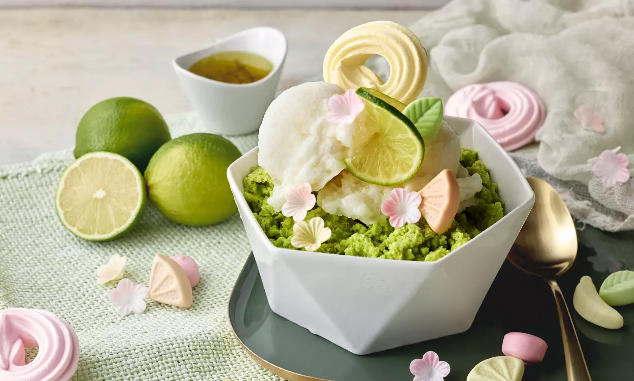 Sorbet cremos alb așezat peste granulat verde intens, garnisit cu felii de lime, frunze delicate și bezele pastel pe o farfurie elegantă