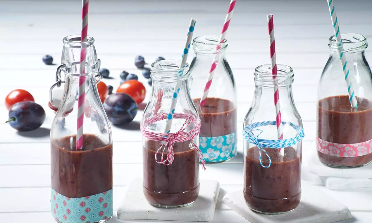 Smoothie dens de roșii roșu-maro servit în sticle transparente decorate cu șnururi colorate și paie, pe fundal cu prune și roșii