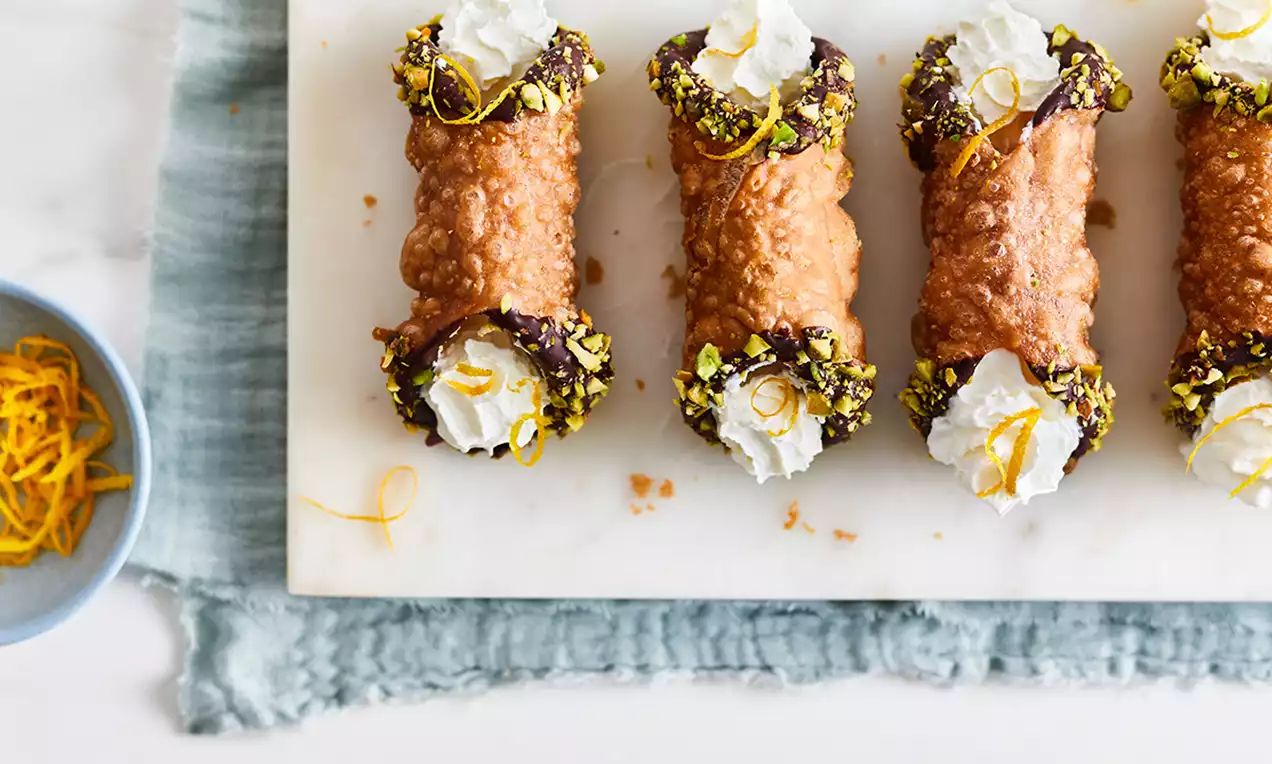 Cannoli vegane cu crustă aurie, capete învelite în ciocolată și fistic, umplute cu cremă albă pufoasă, ornate cu spirale de coajă de portocală
