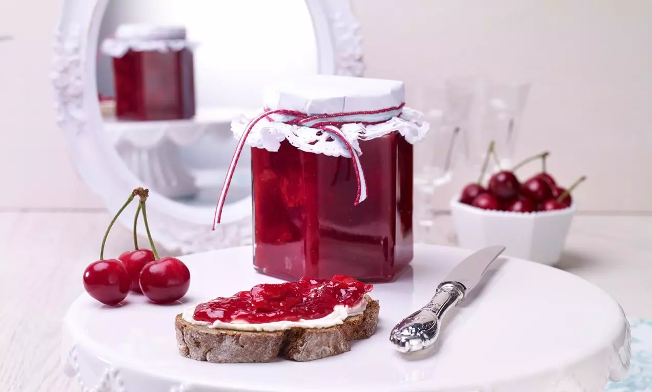 Dulceață de vișine roșu-rubiniu într-un borcan cu capac decorativ, lângă cireșe proaspete și o felie de pâine unsă pe un platou alb