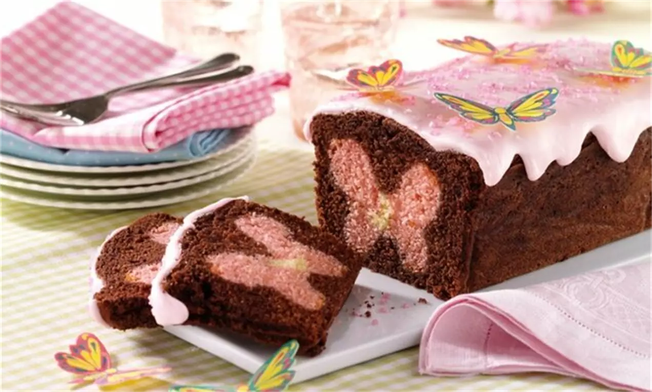 Chec de cacao cu interior surpriză în formă de fluture roz, acoperit cu glazură albă și decorat cu fluturi colorați pe o tavă albă
