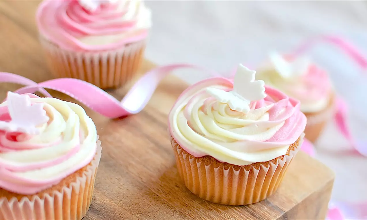 Brioșe aurii cu frosting dublu alb și roz învârtit în forme elegante, decorate cu figurine albe, așezate pe un platou de lemn