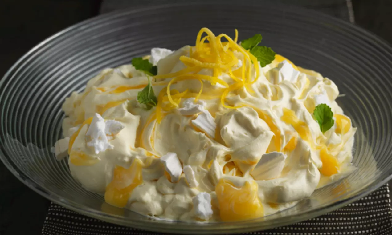 Budincă cremă alb-galbenă cu textură aerată, decorată cu bezea crocantă, coji de lămâie și frunze de mentă pe o farfurie de sticlă transparentă