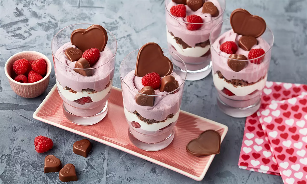 Desert la pahar în straturi de mousse roz și cremă albă, cu zmeură proaspătă și ciocolată în formă de inimă, pe un platou roz