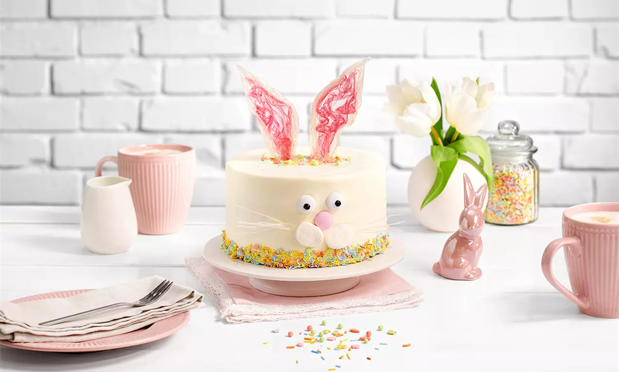 Tort alb-cremos decorat cu urechi roz din zahăr, detalii de iepuraș din marshmallows și ochi dulci, presărat cu bomboane colorate pe margine
