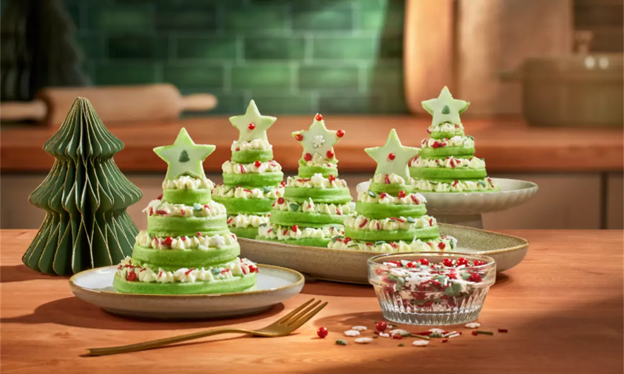 Turnulețe de clătite verzi sub formă de brăduți, decorate cu frișcă, bomboane colorate și stele din biscuiți pe farfurii festive.