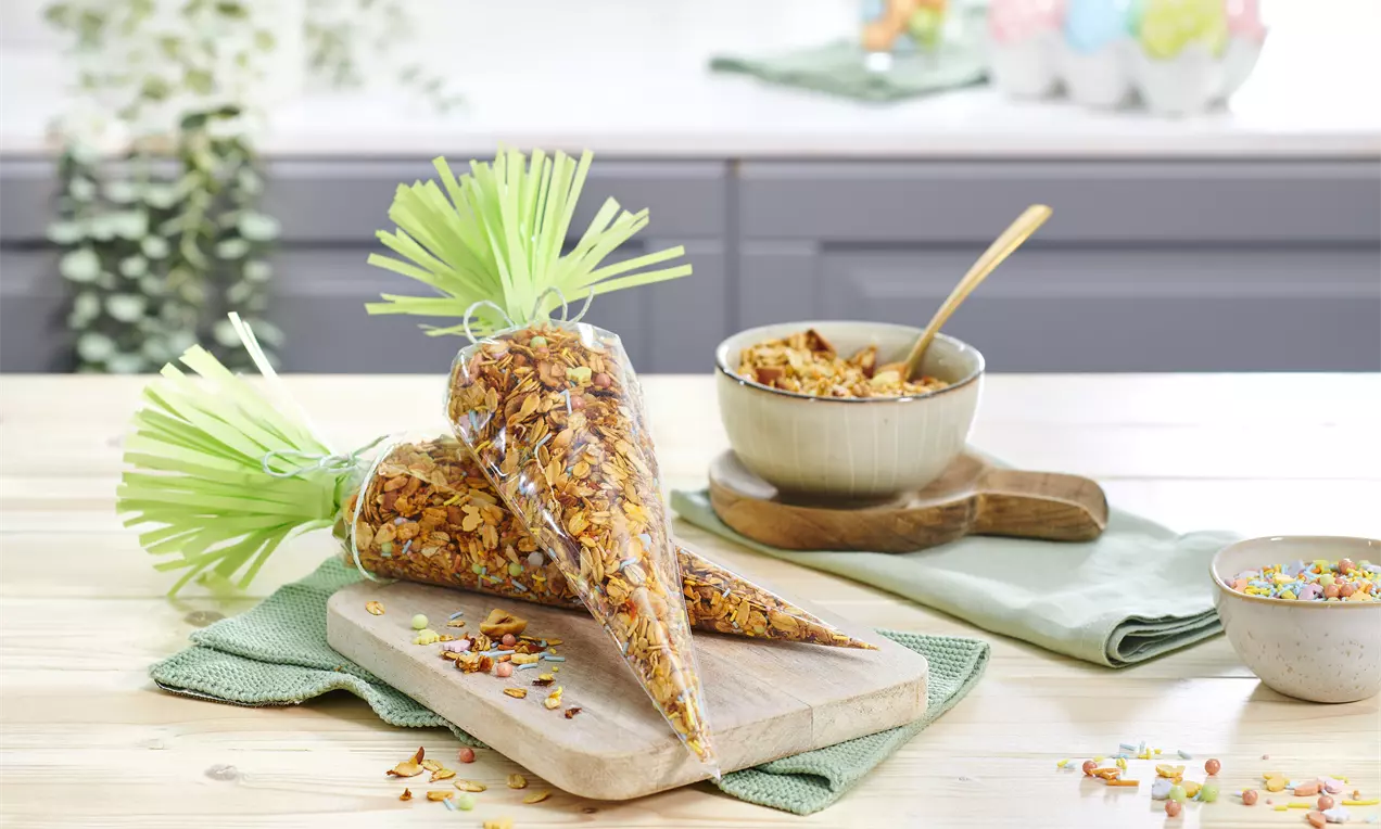 Granola crocantă cu nuanțe arămii, ambalată în pungi conice transparente cu franjuri verzi, așezată pe o masă de lemn deschis la culoare